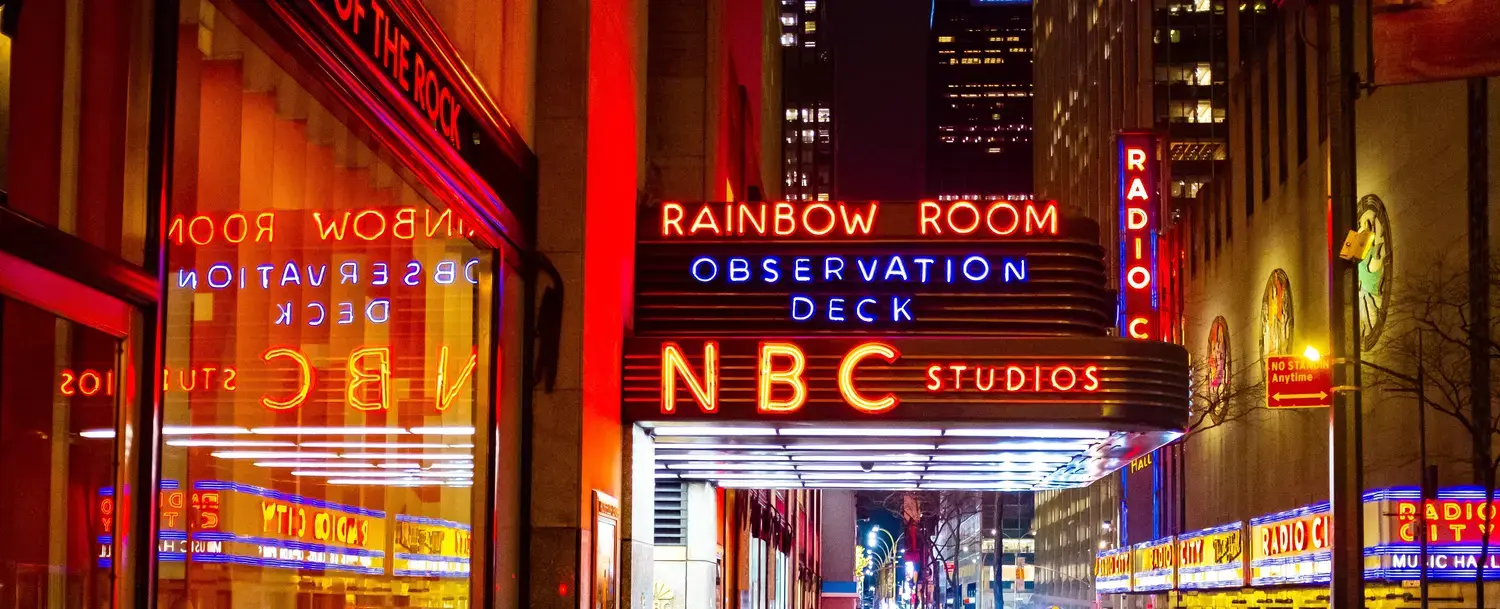 a nighttime view of a building with neon signs reading 'Rainbow Room Observation Deck' and 'NBC Studios'